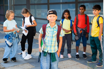 Cute cheerful tween boy looking at camera while standing in school campus with notebooks in hands on background with schoolmates.