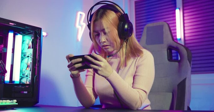 Gamer playing has aches and pains twisting left and right while playing in online video game long times with on smart phone with neon lights, Asian woman wearing gaming headphones play smartphone