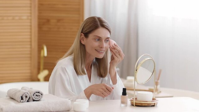 Everyday makeup. Beautiful middle aged lady applying concealer on face with sponge at home, free space