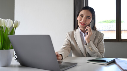 Gorgeous asian woman in business suit talking on mobile phone and using laptop at her office