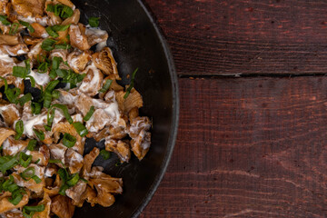 French-fried chanterelle mushrooms with onions and sour cream in a frying pan on a dark wooden background, garlic, onion and parsley and dill greens are laid out next to each other.