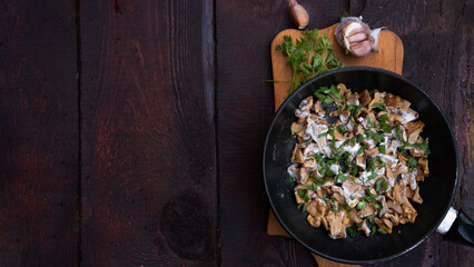 French-fried chanterelle mushrooms with onions and sour cream in a frying pan on a dark wooden background, garlic, onion and parsley and dill greens are laid out next to each other.