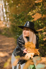 Fototapeta na wymiar Attractive blonde woman in a leather hat and jacket with a bouquet of yellow maple leaves in her hands walks in the autumn park.Autumn concept.Beauty in nature.Slow living.