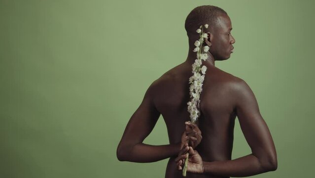 Medium studio portrait of of handsome African American man standing topless holding white flowers behind his back, pale green background