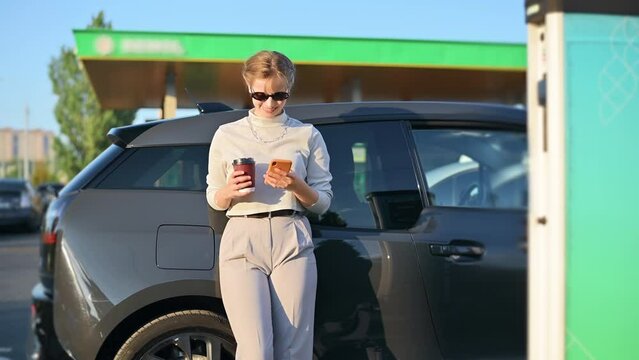 A young blonde woman with smartphone and coffee at a car charging station with electric car nearby in Chisinau, Moldova