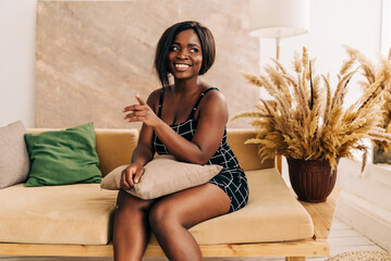 Naklejka premium Happy stylish beautiful female student with hair looks at camera stands in cozy home interior. Smiling attractive young woman wears black dress headshot portrait.