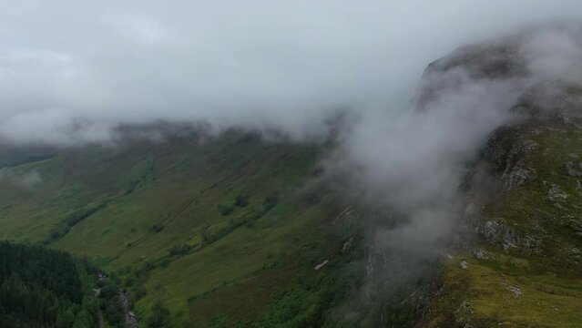 A drone shot in the Scottish highlands coming over a spectacular, cloud covered peak and into a tree covered valley.