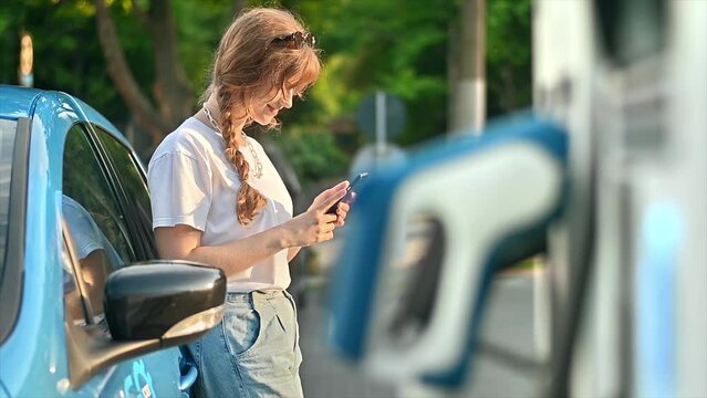 A young blonde woman using smartphone at a car charging station with charging electric car nearby. Chisinau, Moldova. Slow motion