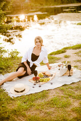 Young attractive girl with dark hair in a white shirt on a picnic in nature by the river in summer at sunset. Healthy life.