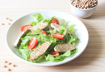 tempeh or tempe salad with tomato and green vegetable in white plate and a bowl of soy beans on wooden table.