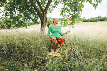 Obraz premium Middle aged woman with short light hair, green top and colorful flower pants in high meadow holding colorful pinwheel