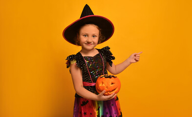 Little cute girl wearing witch costume, holding orange pumpkin with toy spider, pointing index...