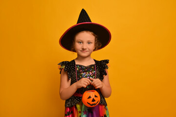 Little cute girl in witch costume holding Halloween pumpkin candy bucket or Jack o Lantern candy holder and looking at camera on a yellow background.

Happy Halloween, masquerade or party concept.