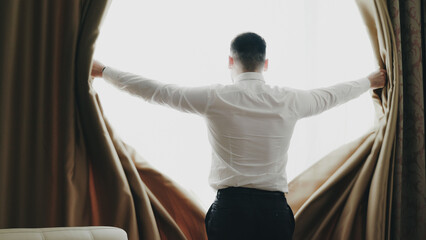 Businessman open curtains in hotel room at the morning and looking into window