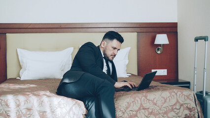Fototapeta premium Pan shot of concentrated young businessman in suit working on laptop while sitting on bed in hotel room. Business, travel and people concept