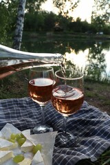 Pouring delicious rose wine into glass on picnic blanket near lake, closeup