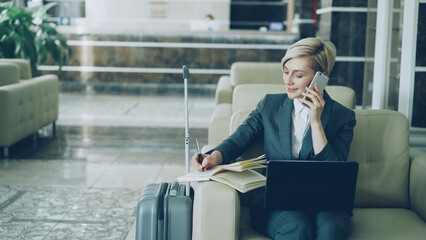Pan shot of blonde busy businesswoman sitting in armchair in hotel lobby talking mobile phone,...
