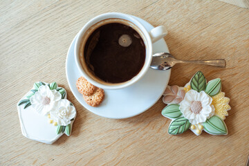 Fototapeta na wymiar Cup of hot flavored black coffee with soft foam on saucer and nice icing flower shaped cookies on table top view closeup, free copy space. Catering, coffee house, serving food, drink and dessert
