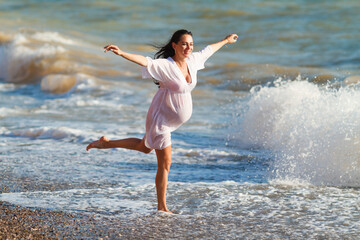 Pregnant woman is jumping on beach. Cheerful pregnant woman runs on seashore. Pregnant woman in a romantic white dress jumping on the beach near the tropical sea. Indication of happy pregnant women.