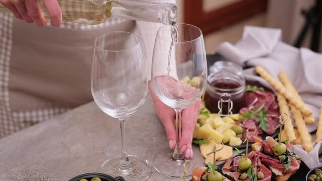 woman pouring wine to a glass from a bottle at domestic kitchen with meat and cheese plater on the table