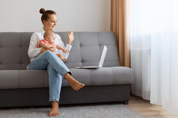 Indoor shot of smiling woman in white t shirt sitting on sofa with baby daughter and working on...