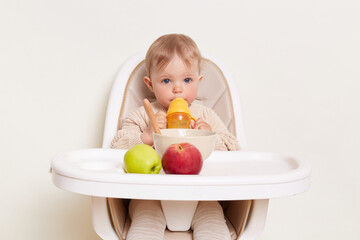 Indoor shot of cute thirsty baby wearing beige sweater sitting in a child's chair and drinking...