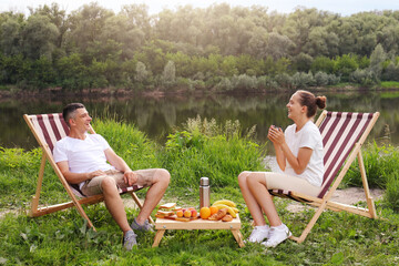 Image of happy delighted man and woman sitting on deck chair and drinking coffee or tea near the...