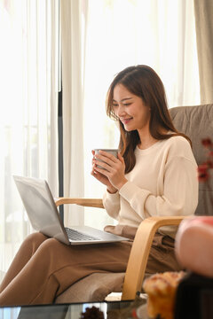 Beautiful woman drinking tea and checking email on her laptop, relaxing at home in a bright winter morning