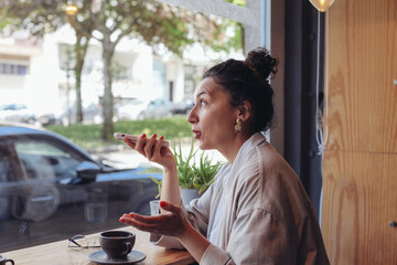 A woman recording a audio message with a smartphone. A woman sitting in a cafe by the window with a cup of coffee
