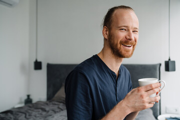 Young ginger man drinking coffee while sitting on bed