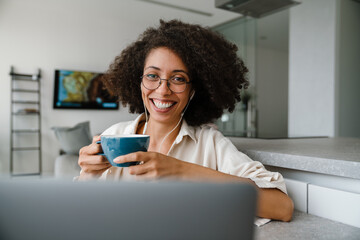 Black woman in earphones drinking coffee while working with laptop