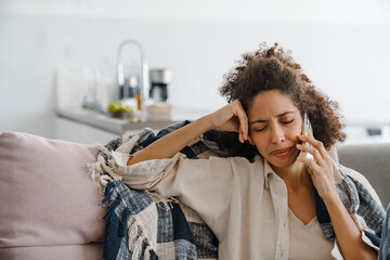 Black displeased woman talking on mobile phone while sitting on sofa