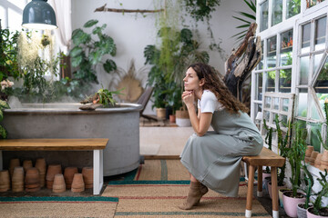 Sad pensive woman sits on bench in home garden, thinking of crisis challenges looking out the window. Thoughtful gardener female resting after work in floral store or greenhouse. Doubtful lady at home