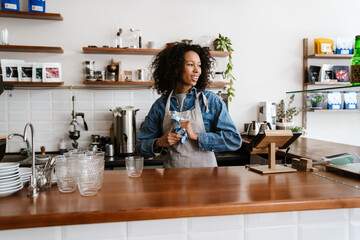 Black barista woman wearing apron wiping glasses while working in cafe