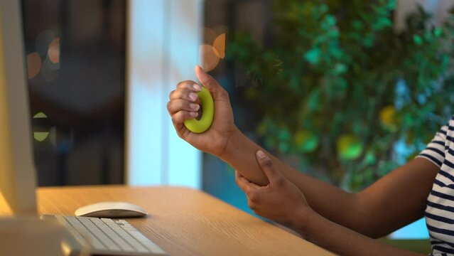 African American woman squeezing rubber expander during working day in office, employee using hand exerciser for carpal tunnel syndrome prevention while working on computer. Stress relief at work.