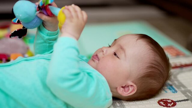Adorable healthy Caucasian kid in blue clothes lies on his back. Active baby waving his toy in the air. Close up. Blurred backdrop.