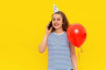 Fascinating happy young woman is talking on a mobile phone, receives congratulations from friends on anniversary, wears party cone and striped tshirt, feels very happy, standing over yellow background