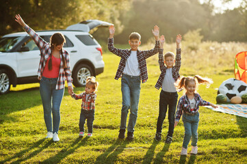Family spending time together. Mother with four kids standing and holding hands against white suv car. - obrazy, fototapety, plakaty