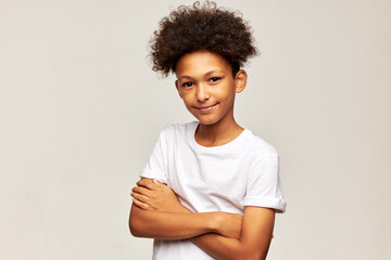 Fototapeta premium Portrait of cute adorable african american boy with afro hairstyle standing on gray background with crossed arms looking at camera smiling. Childhood, body language, sign, gestures concept