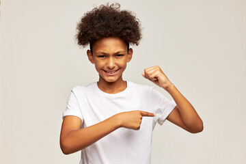 Studio portrait of strong teen kid with afro hairstyle posing on gray background in white shirt showing his biceps muscles with furious facial expression, ready to fight, to protect himself