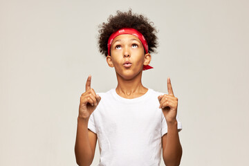 Amazed surprised african american teen boy in red bandana and white mockup t-shit with copy space for your advertising content pointing fingers up, saying ooh wow on gray studio background