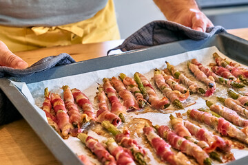Man blogger preparing young green asparagus sprouts wrapped in bacon on wooden table in the kitchen. Hands of man holding baking tray with asparagus wrapped in ham baked in oven.