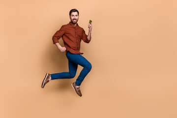 Fototapeta na wymiar Full body profile photo of active cheerful guy jump run hurry hold telephone isolated on beige color background