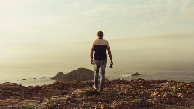 Travel photographer walking towards the cliff to take a panoramic landscape photo at sunset. Travel photographer from behind with camera in hand looking at an island on the horizon