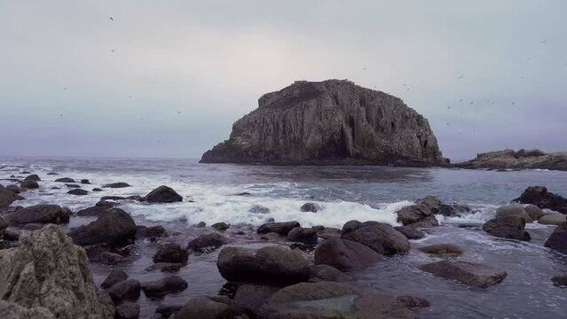 Low angle of sea hitting rocky shore near flock of birds resting in Pe&ntilde;a Blanca rocky islet, Algarrobo, Chile