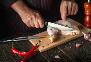 Preparing for cooking chicken legs in the kitchen. Chef cuts a raw chicken leg with a knife on a...