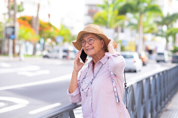 Cheerful attractive senior woman with summer hat standing in the city street using mobile phone