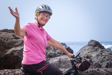 Active happy senior woman with helmet running on her electric bicycle along the sea looking at...