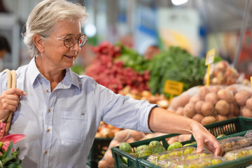 Senior woman at the farmers market holding a shopping bag looking at fruits and vegetables, price increase and inflation
