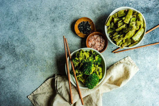 Overhead view of bowls of green beans and broccoli with sesame seeds and pink himalayan salt